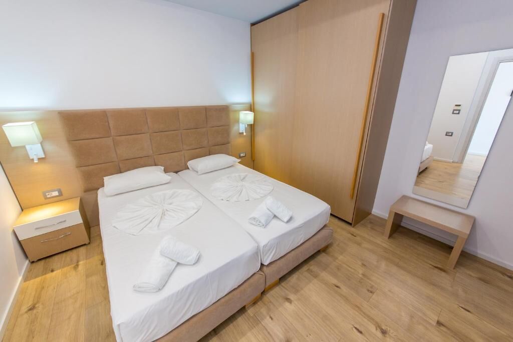 A double bed with white linens and towels, a wooden headboard, bedside tables with lamps, and a wooden bench.