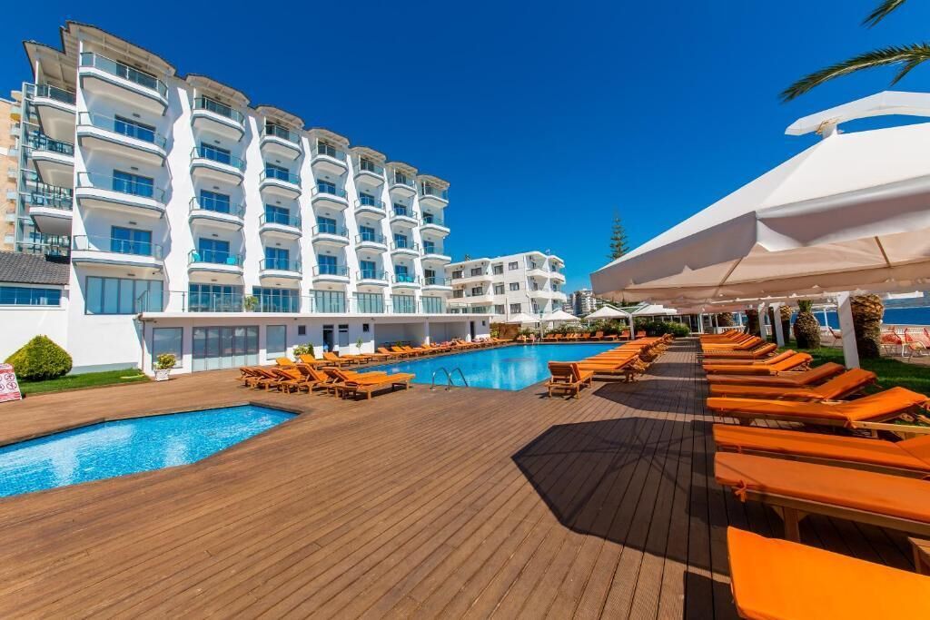 A poolside area with lounge chairs and a swimming pool, surrounded by a wooden deck and white buildings with balconies.
