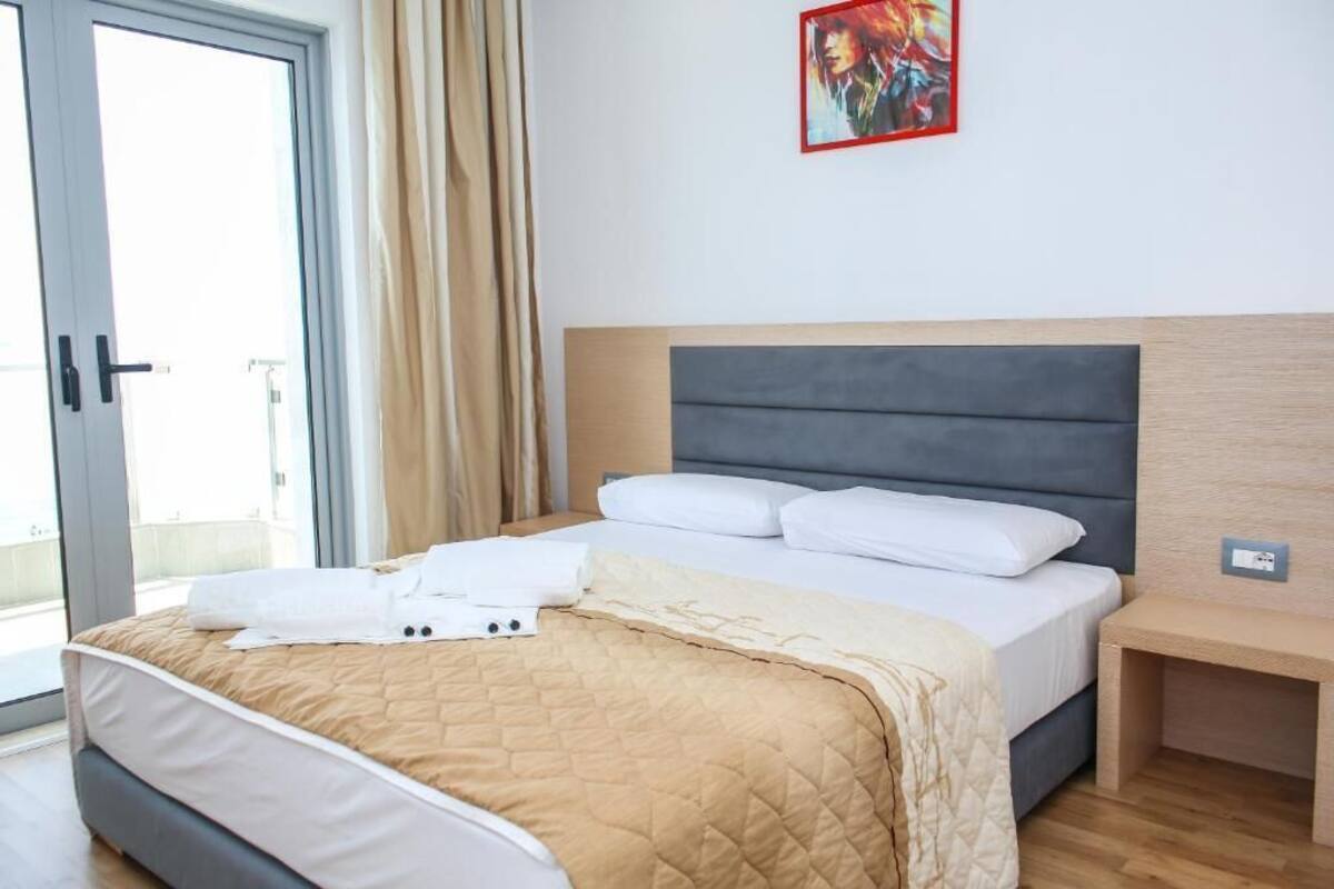 A hotel room with a bed, a bedside table, a window with curtains, and a painting on the wall.