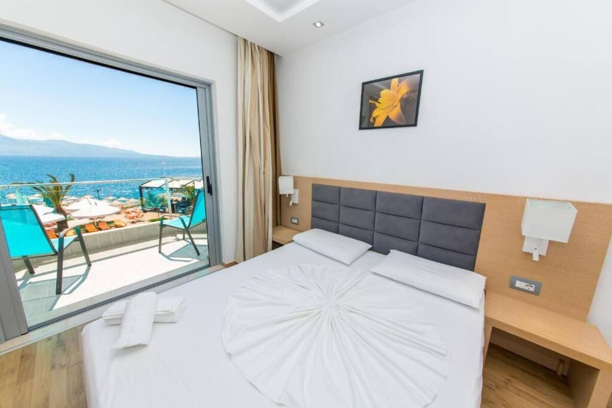 A hotel room with a bed, a nightstand, a bedside lamp, a window with a view of the sea, and a chair by the window.