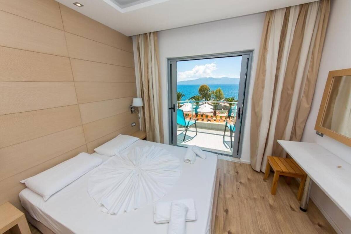 A hotel room with a bed, a wooden nightstand, a small table, and a view of the sea through a glass door.