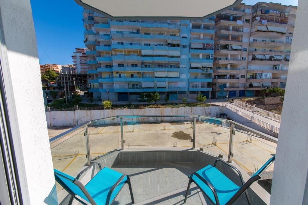 A rooftop terrace with two blue chairs, a glass balustrade, and a view of a multi-story residential building.