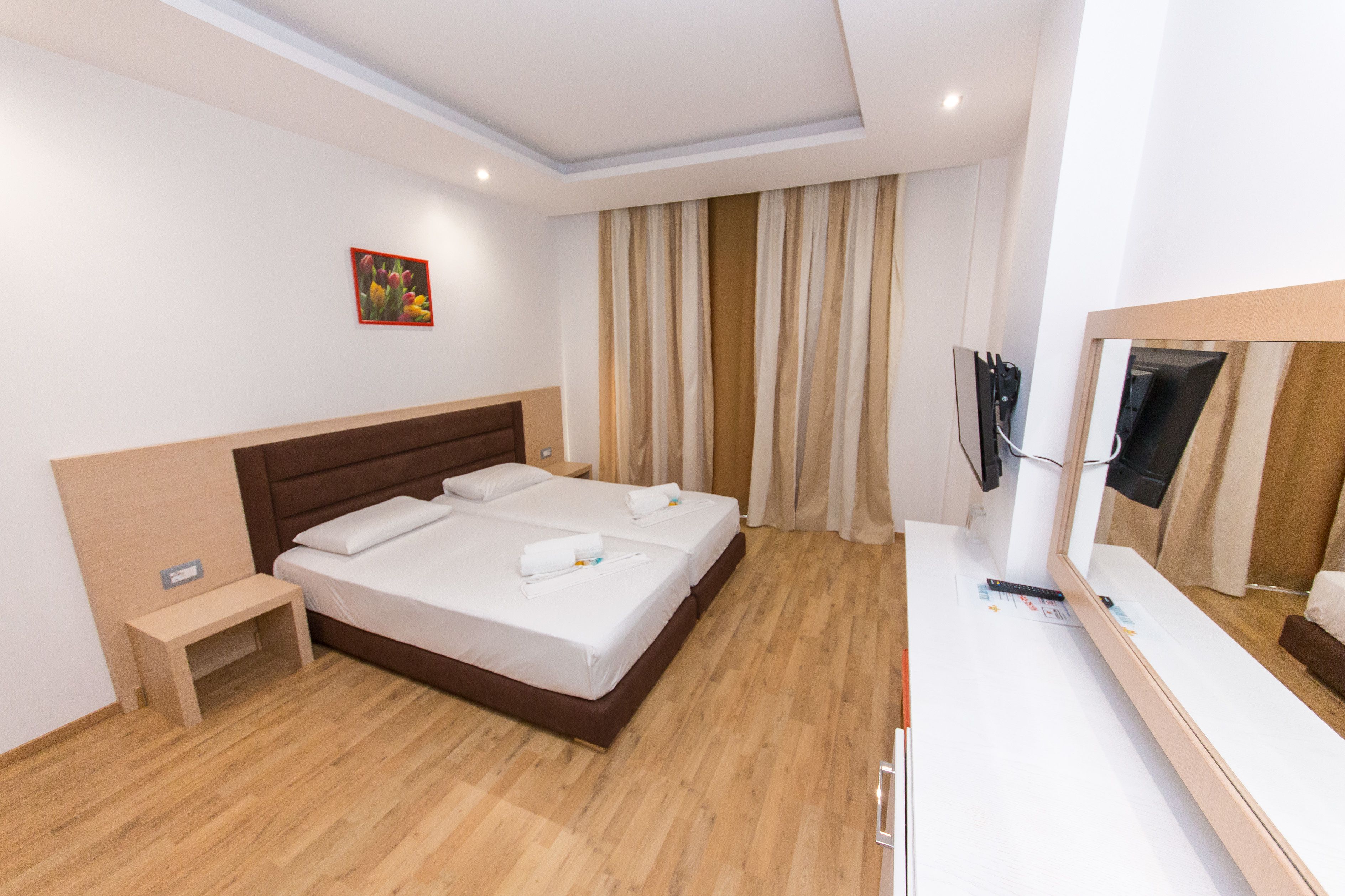 A hotel room with a double bed, a bedside table, a small bench, a mirror, and a television.