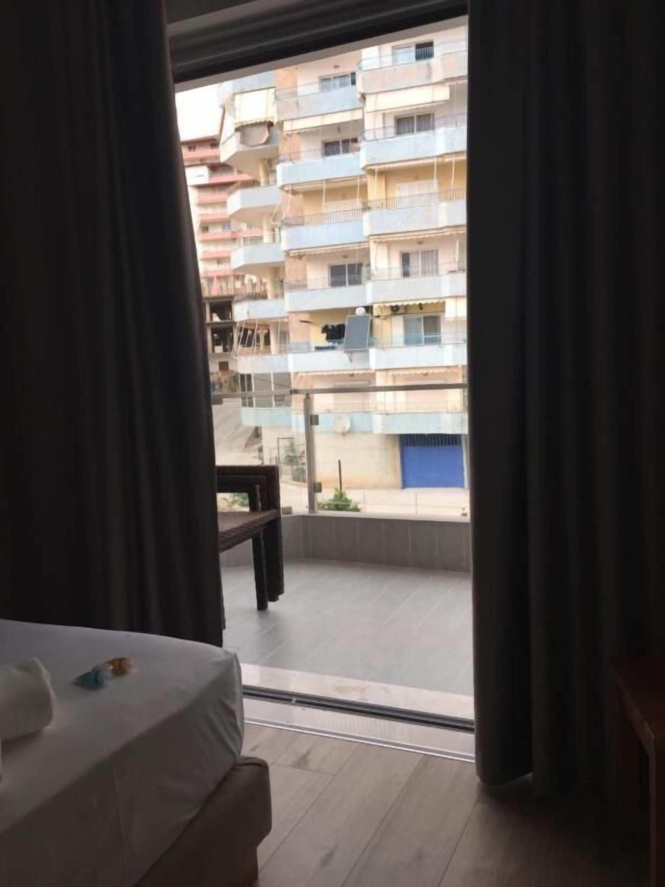 A hotel room with a bed, a small table, and a view of a multi-story building with balconies.