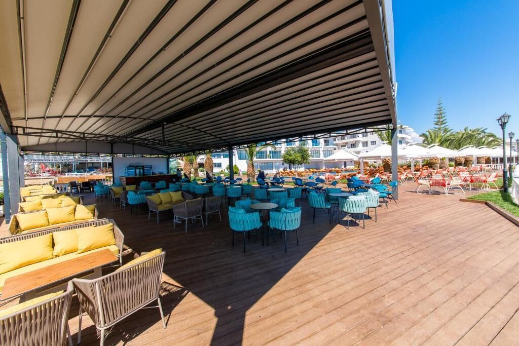 An outdoor seating area with wooden tables and chairs, a covered section with a striped roof, and a view of a building with white umbrellas in the background.