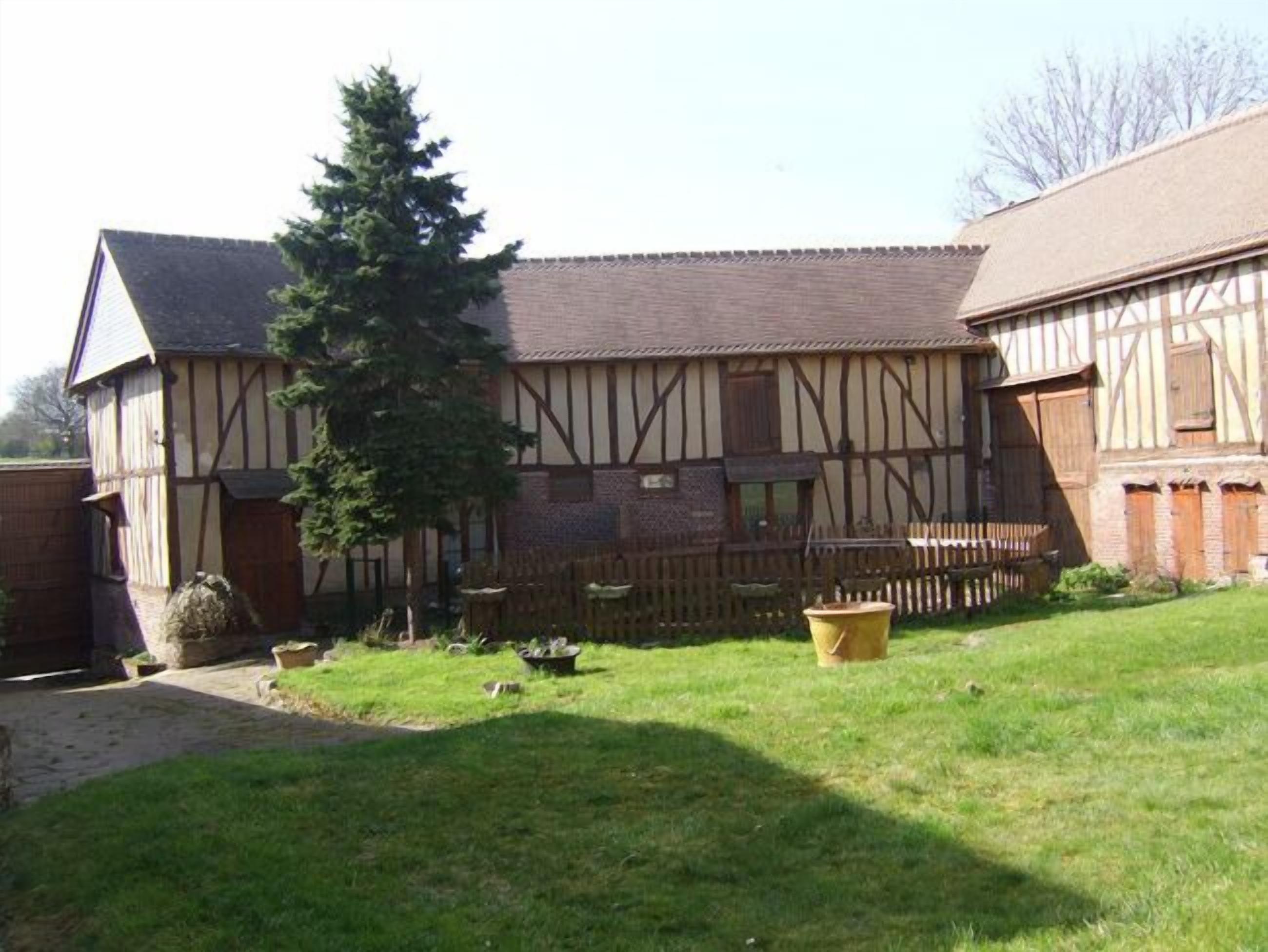 Il s’agit d’un bâtiment traditionnel à colombages, doté d’un toit en pente, entouré d’une cour clôturée où se trouvent un grand arbre et un tonneau jaune.
