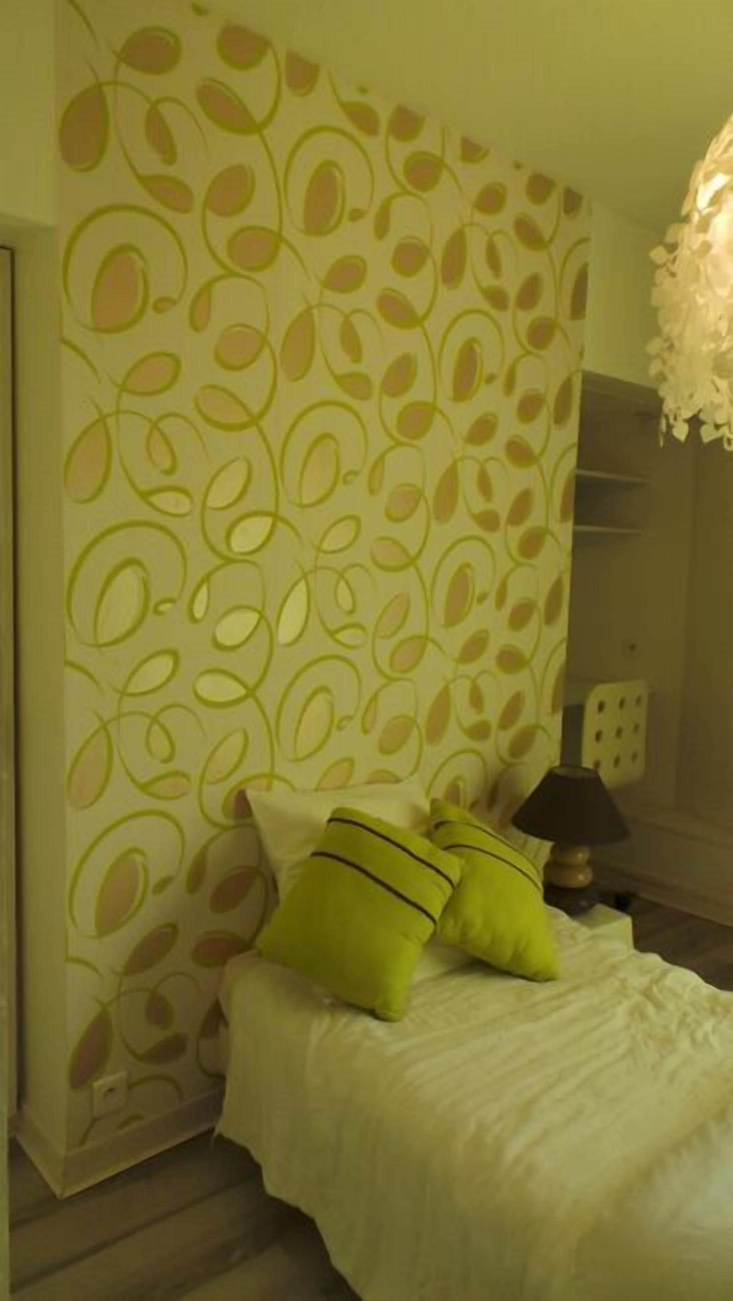 Un dormitorio con un cabecero estampado, una cama con ropa de cama blanca y almohadas verdes, una lámpara de noche y un enchufe de pared.