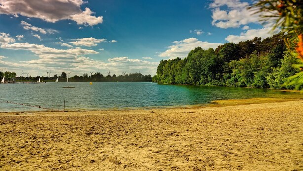 A beach with a sandy shore, a calm body of water, and a forested area in the background. - Campingpark Heidewald (Sassenberg)