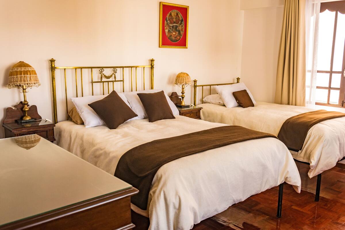 A hotel room with two beds, wooden headboards, bedside tables with lamps, a framed artwork on the wall, and a window with curtains.
