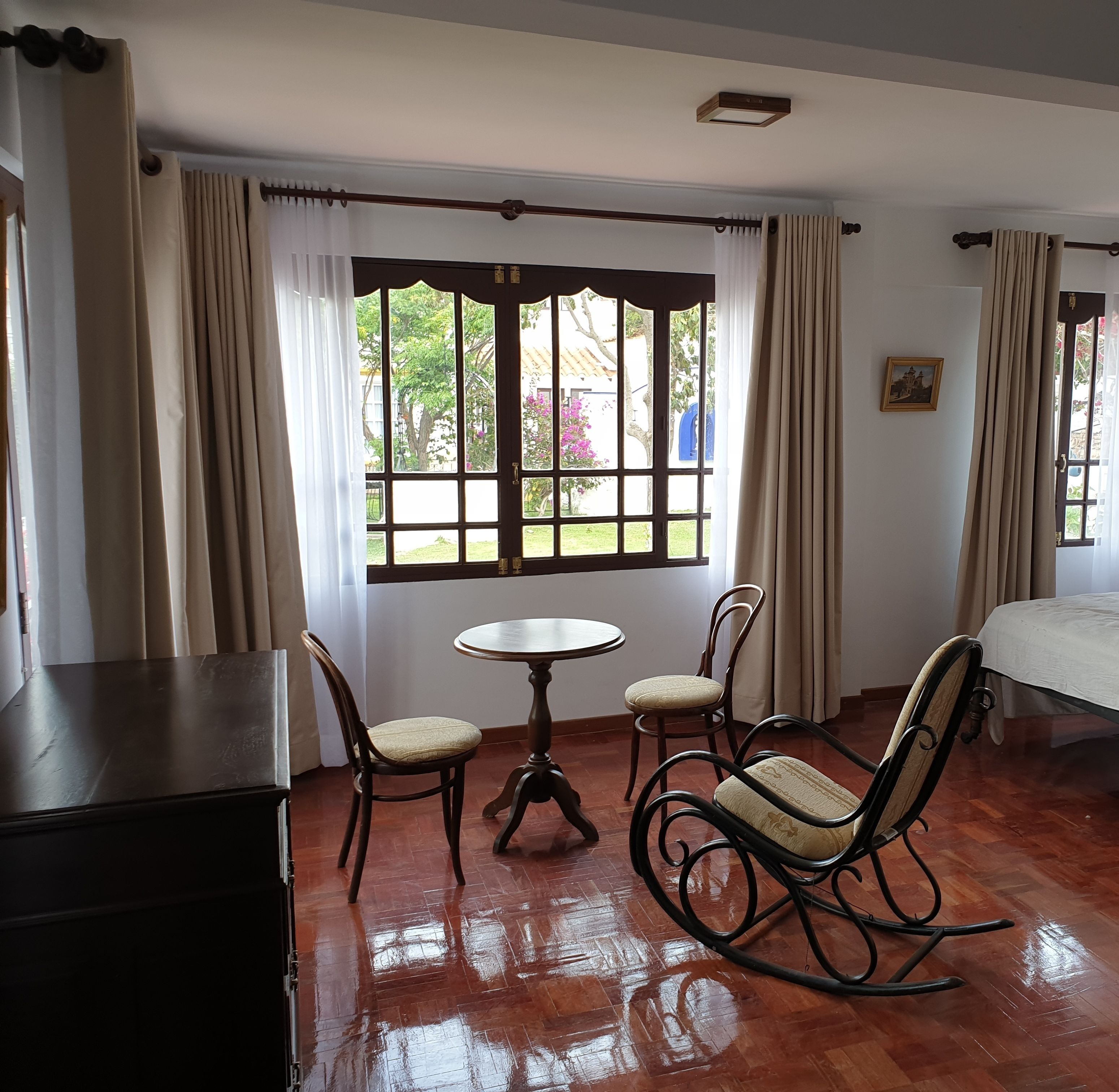 A room with a wooden floor, a round table with two chairs, a bed, and a window with curtains.