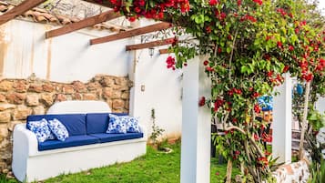 Un área de estar exterior con muebles blancos, un sofá azul y cojines estampados, una pared de piedra y una pérgola con flores rojas.