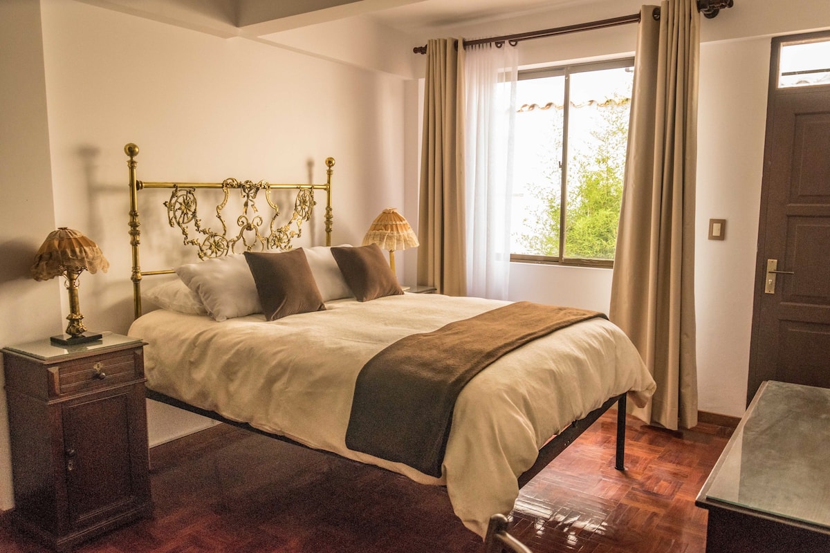 A bedroom with a large bed, two bedside lamps, a wooden nightstand, and a window with curtains.