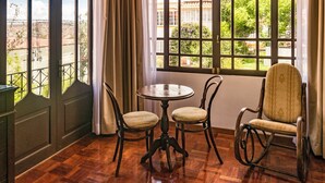 A room with a round table and four chairs, large windows with curtains, and a view of greenery and buildings outside. - El Jardin De Su Merced B&B (Sucre)
