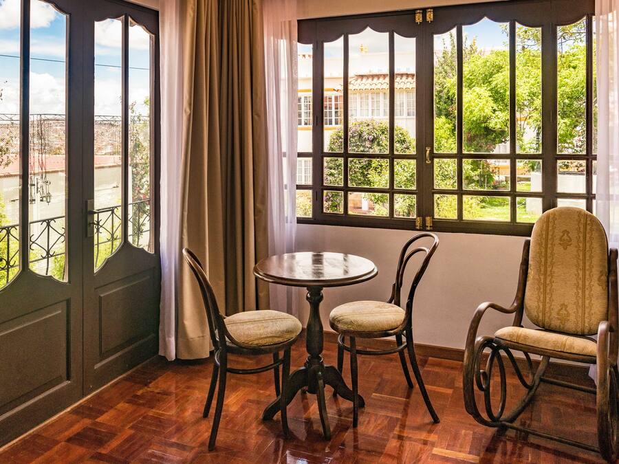 A room with a round table and four chairs, large windows with curtains, and a view of greenery and buildings outside.