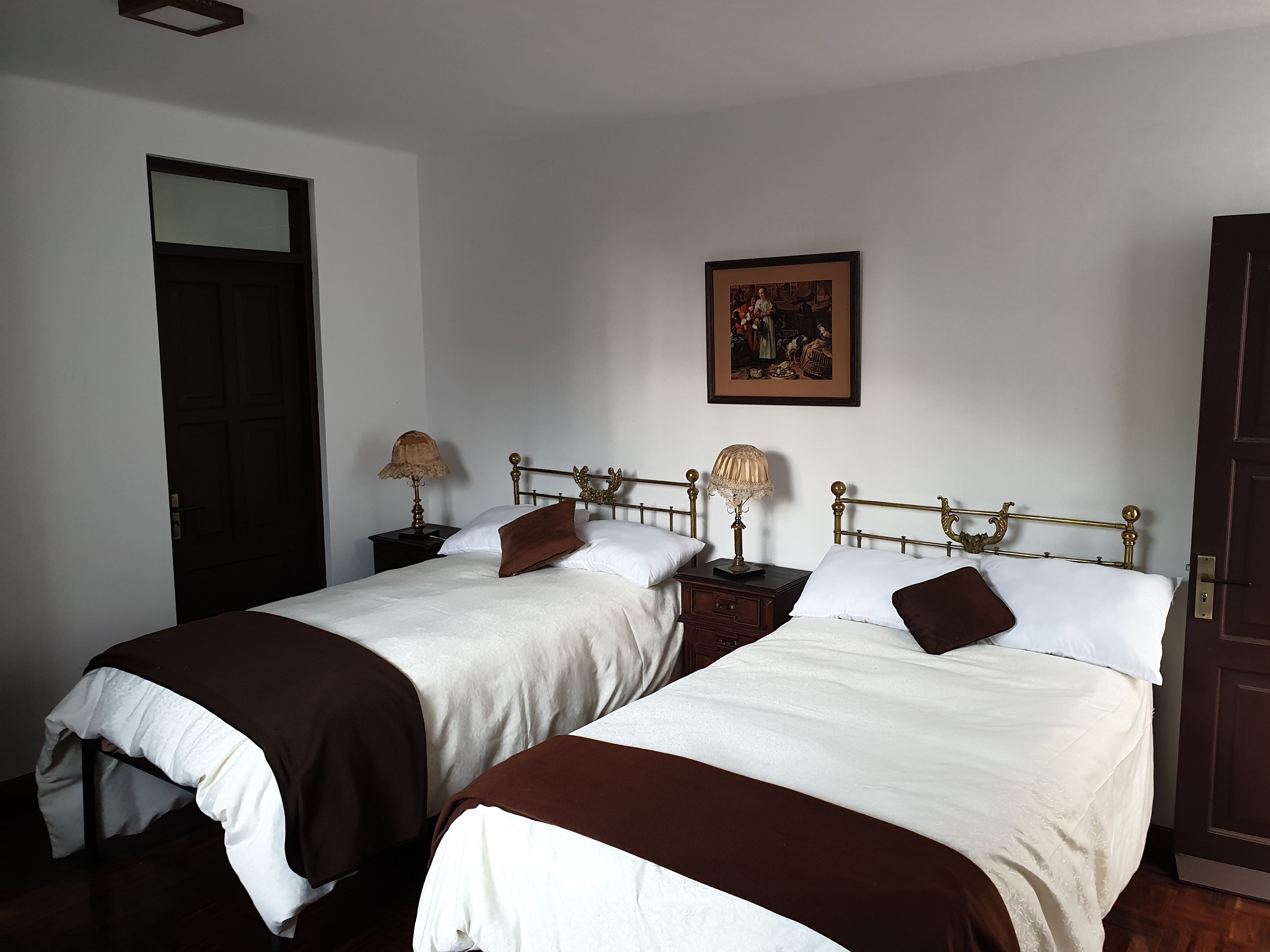 Une chambre avec deux lits, chacun doté d’une tête de lit en bois, de draps blancs et de plaids marron. On y trouve des tables de chevet avec des lampes et un tableau encadré au mur.