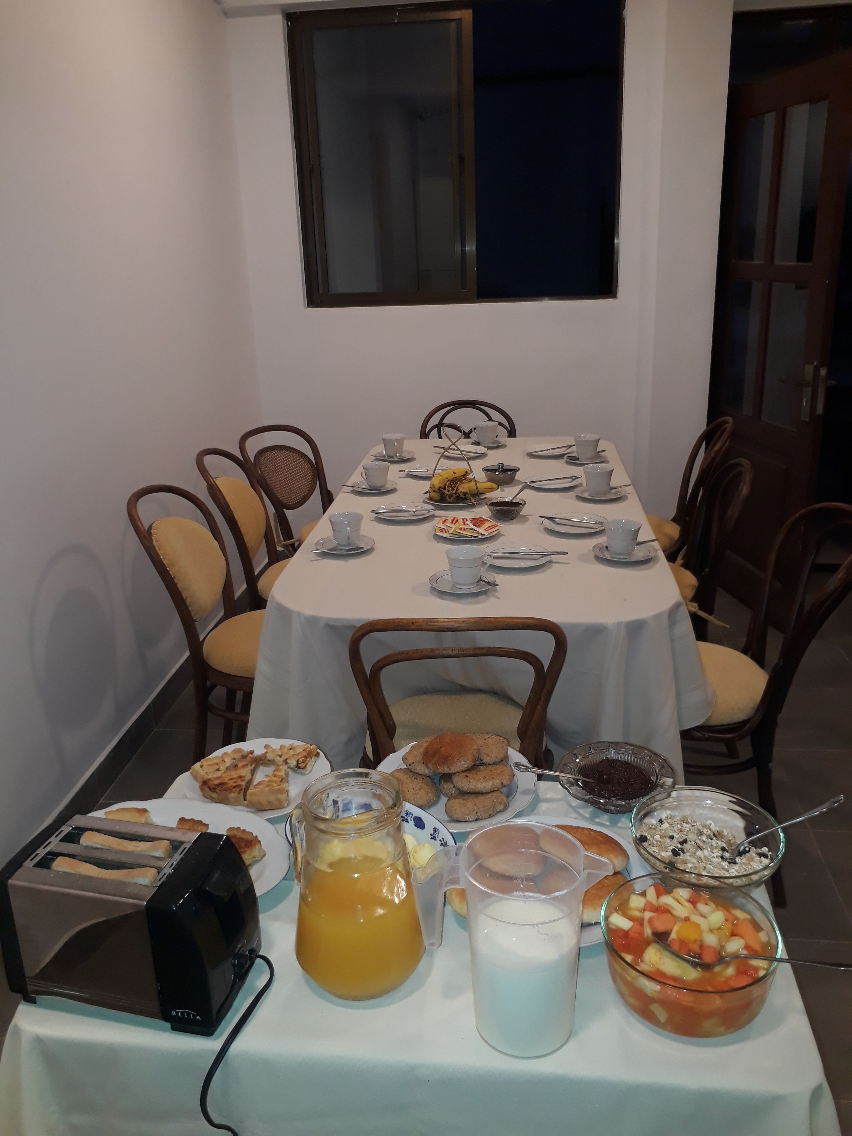 Une table à manger dressée pour un repas, avec différents plats, un grille-pain et une carafe de jus.