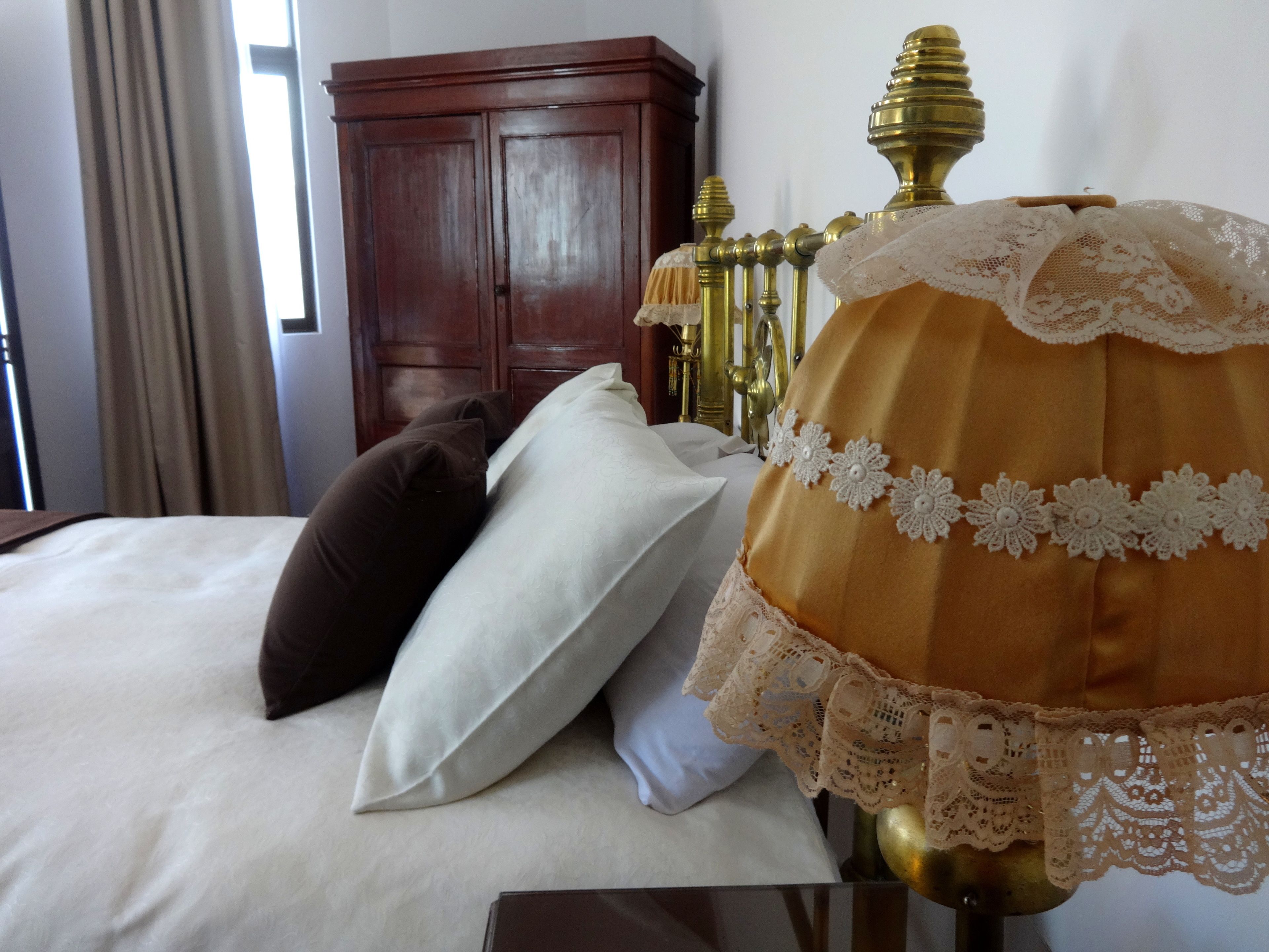 A bed with white and brown pillows, a brass lamp, and a wooden wardrobe in a hotel room.