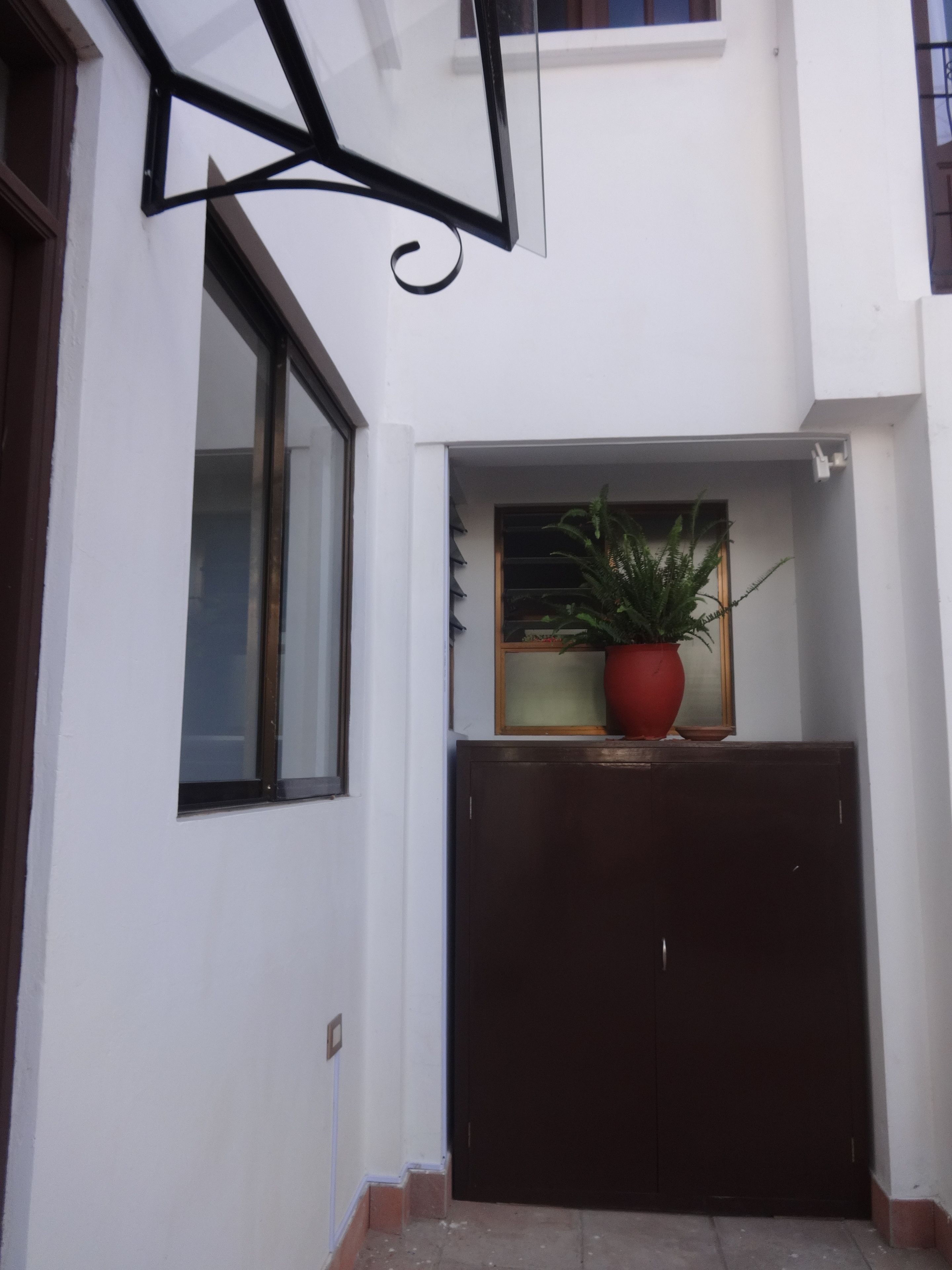 A building entrance with a dark wooden door, a potted plant on a shelf, and a window with blinds.