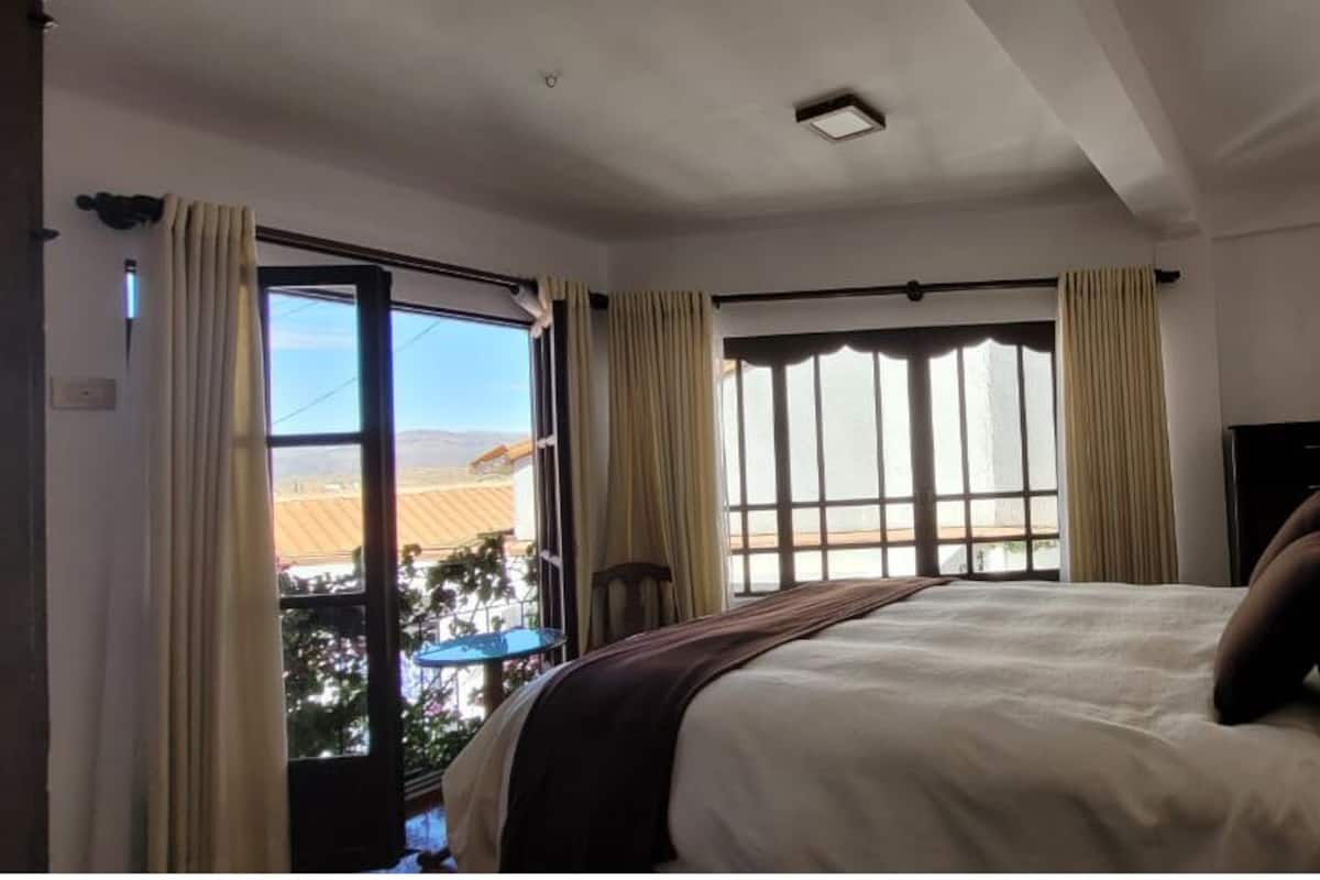 A bedroom with a bed, a window with curtains, a lamp, and a view of the outside.