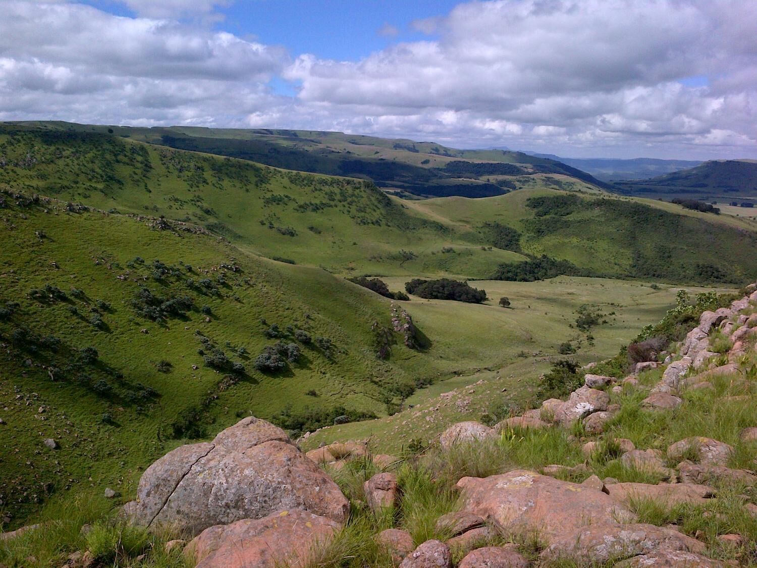 A panoramic view of a green valley with rolling hills and scattered rocks.