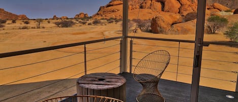A balcony with two wicker chairs and a round wooden table, overlooking a desert landscape with red rock formations.