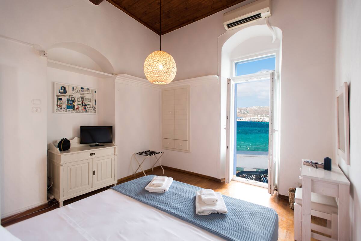 A bedroom with a bed, a TV on a cabinet, a towel rack, and a view of the sea through an open door.