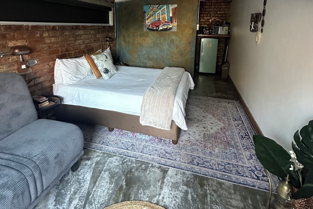 A compact room with a bed, a sofa, a small kitchenette, and a brick wall.