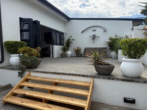 A modern outdoor area with a wooden bench, potted plants, and a white wall with a unique design element. - Relaxed City Living (Gqeberha (Port Elizabeth))
