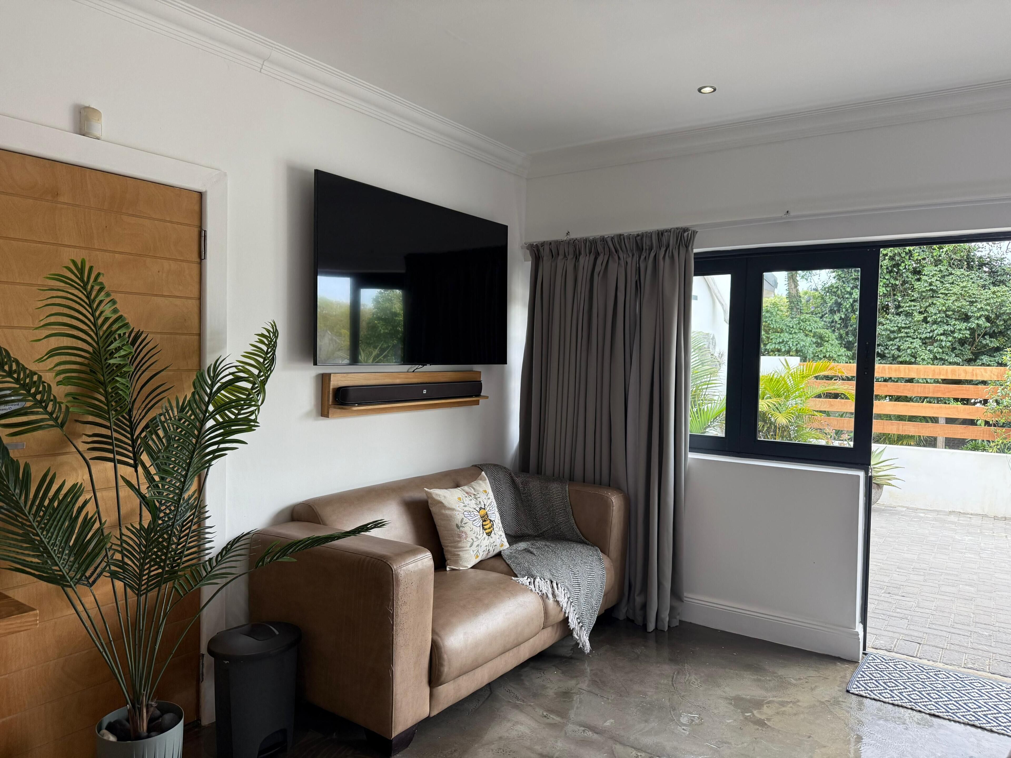 A modern living room with a leather sofa, a mounted flat-screen TV, a large potted plant, and a sliding glass door leading to an outdoor area.