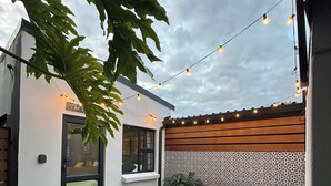 Un patio con mesa y bancos de madera, luces colgantes y un muro decorado.