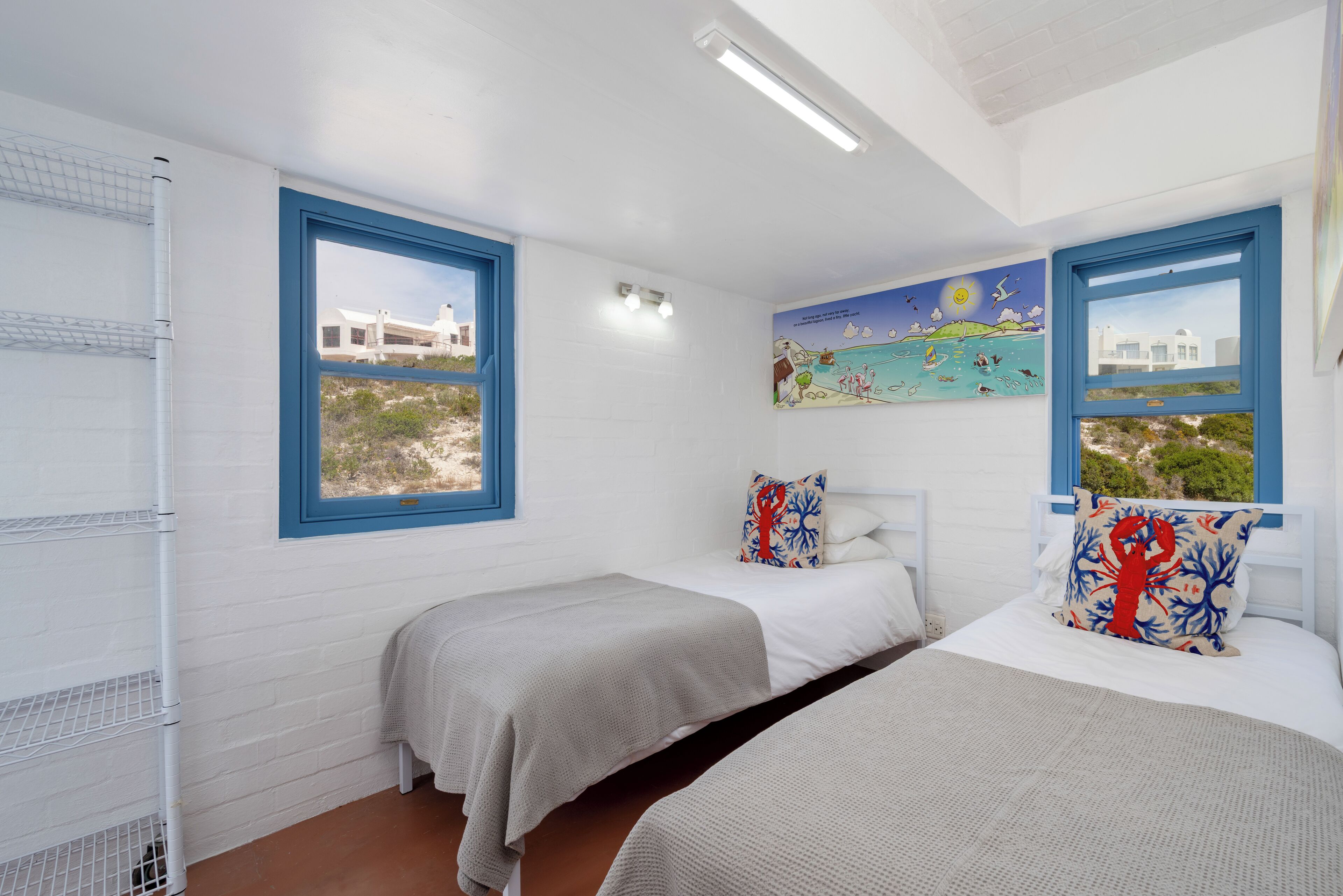 Quarto com duas camas, paredes brancas, molduras de janela azuis, um mural com tema praiano e vista para um edifício.