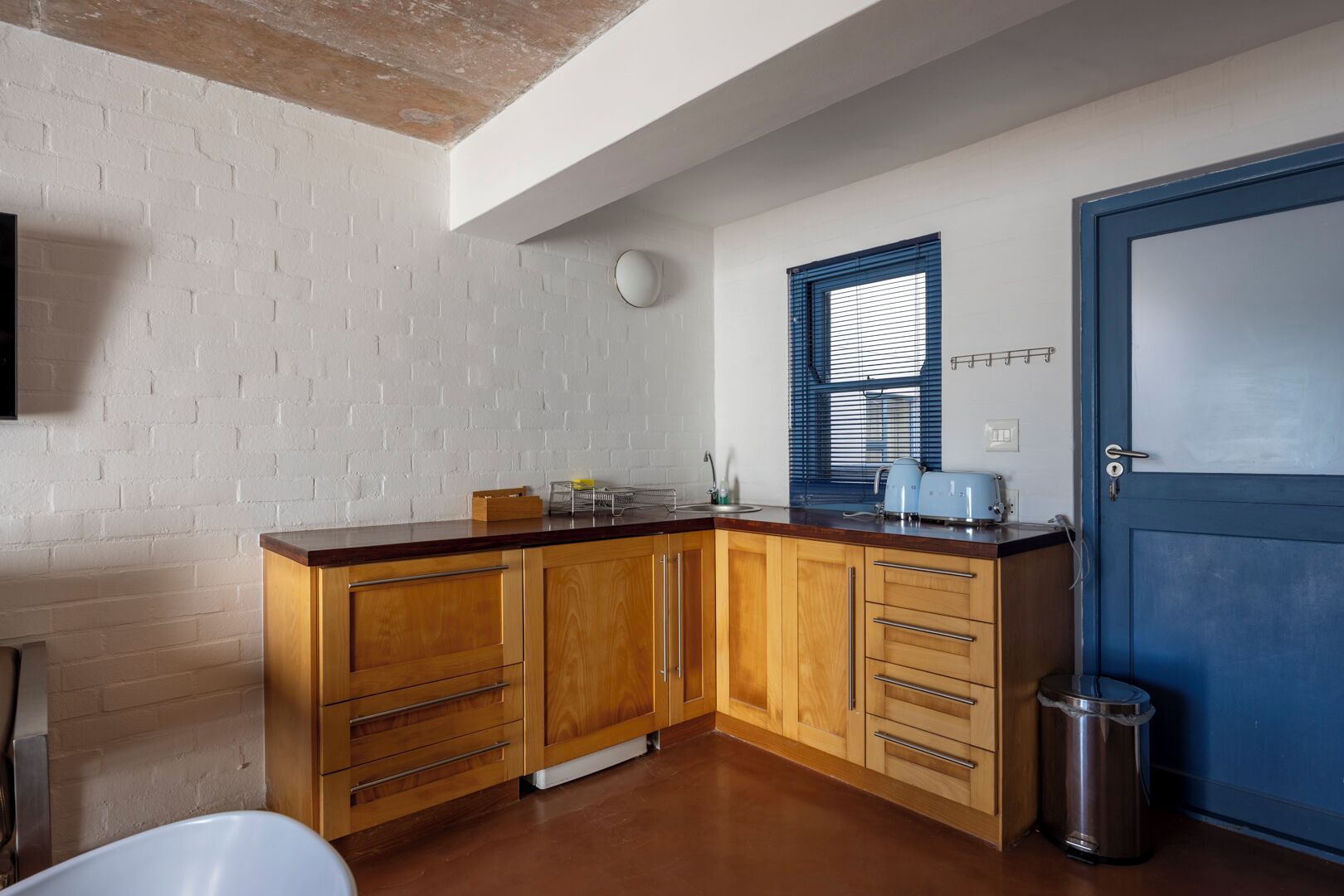 A kitchen with wooden cabinets, a blue door, a window with blinds, and a trash can.