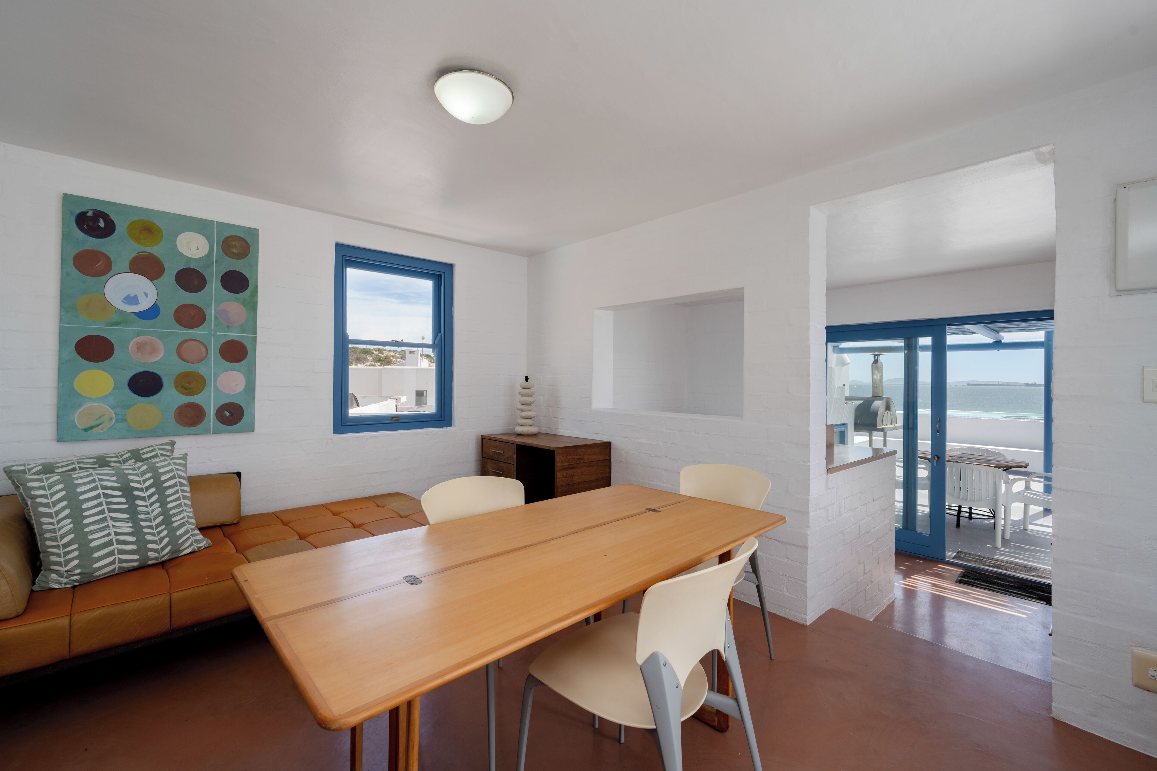 A modern living room with a wooden table and chairs, a sofa with cushions, a colorful abstract painting, a wooden cabinet, and a view of the sea through a door.