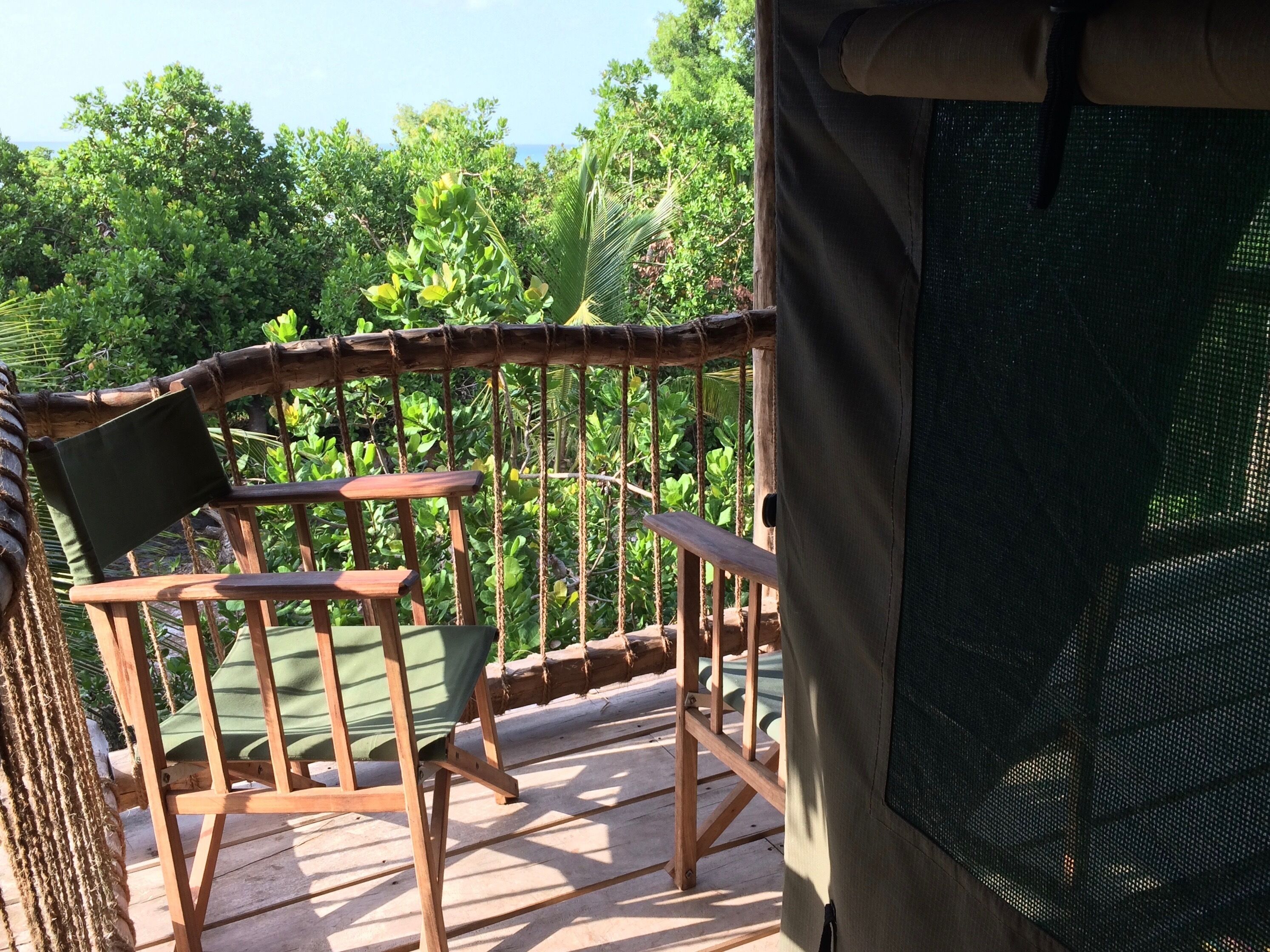 A wooden deck with a chair and a small table, surrounded by lush greenery and a view of the ocean.