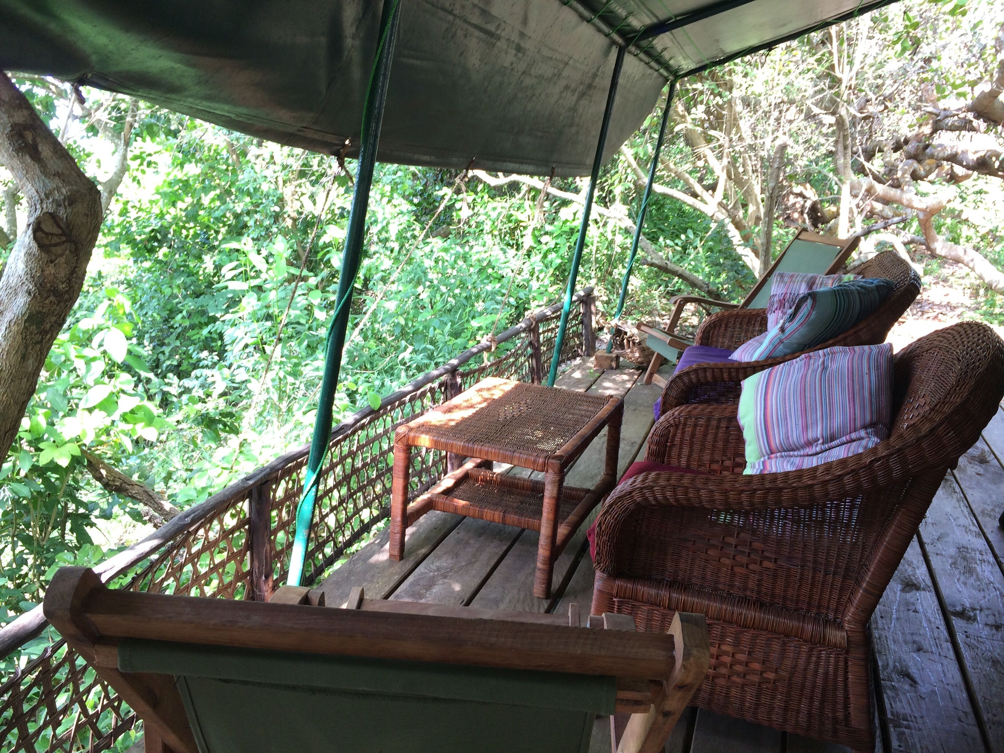 A covered wooden deck with wicker furniture and a view of a forested area.