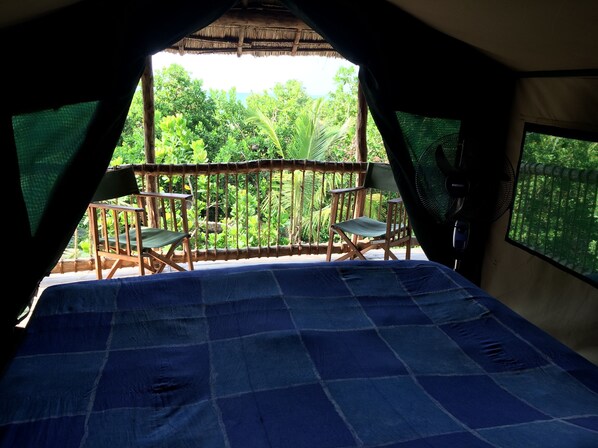 A room with a bed, two chairs, a fan, and a view of lush greenery outside.