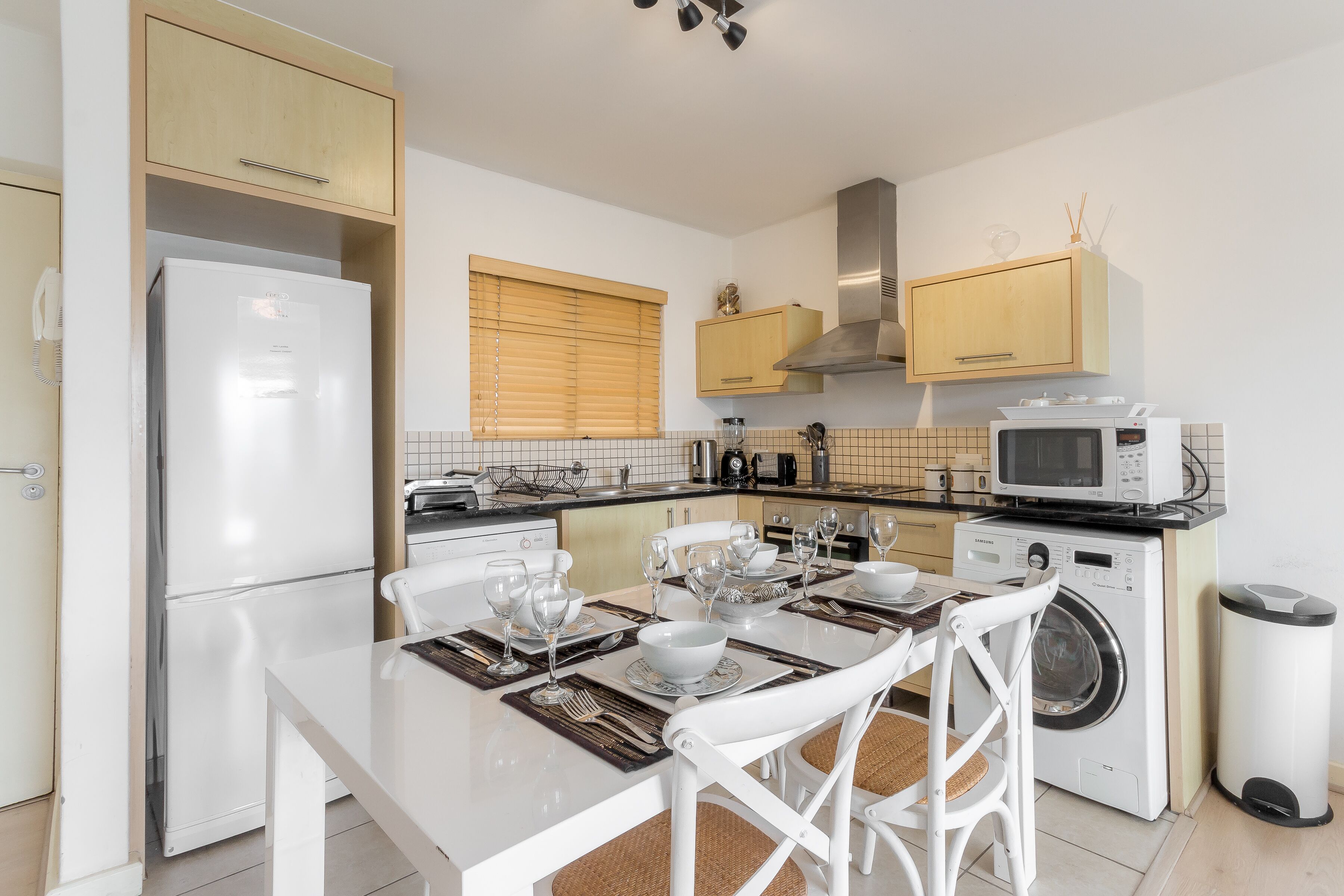 Une cuisine moderne avec un réfrigérateur blanc, un four à micro-ondes et un lave-linge. On y trouve une table à manger mise avec des assiettes, des verres et des couverts.