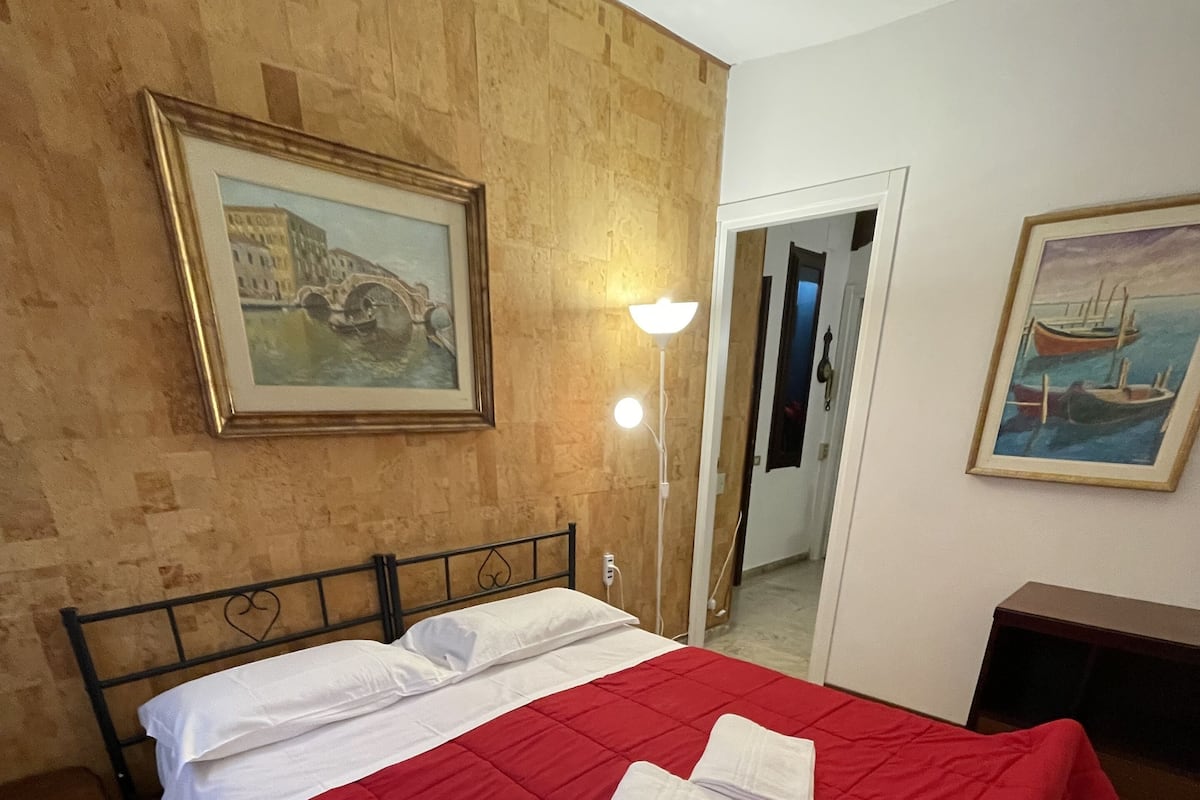 A hotel room with a bed, a red bedspread, two white towels, a nightstand, a lamp, and two paintings on the walls.