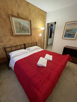 A hotel room with a bed, a red bedspread, two white towels, a nightstand, a lamp, and two paintings on the walls. - Royal Guest House Venice (Venice)