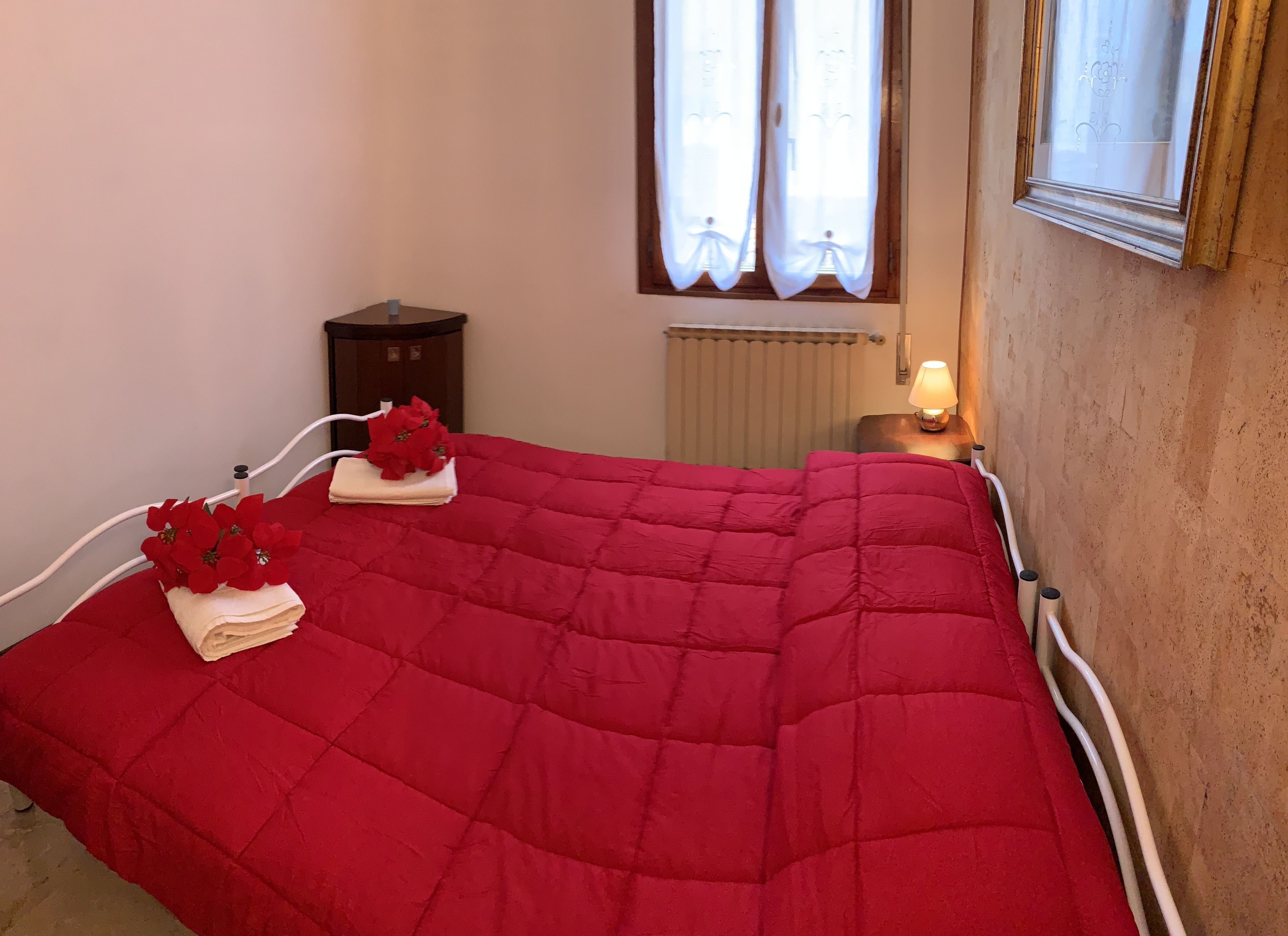A neatly made bed with a red quilt, a small bedside table with a lamp, and a window with curtains.