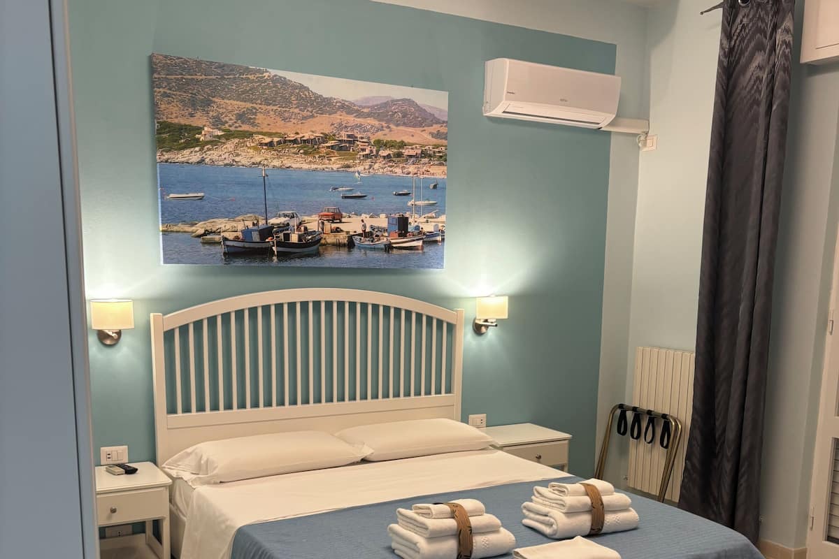 A neatly arranged bedroom with a bed, bedside tables, a wall-mounted air conditioner, and a painting of a coastal scene.