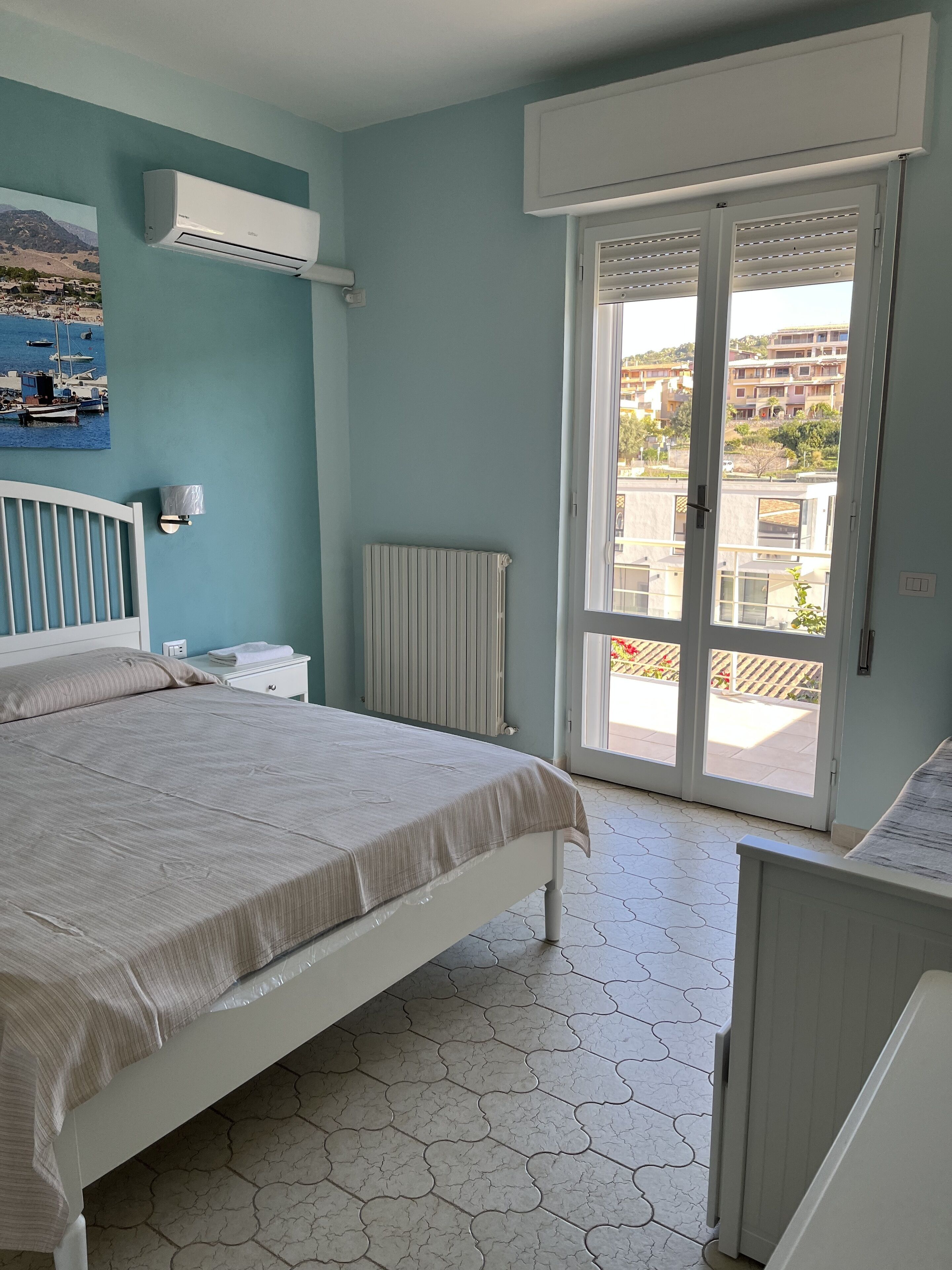 A bedroom with a bed, a radiator, a window with a view of buildings, and a wall-mounted air conditioner.