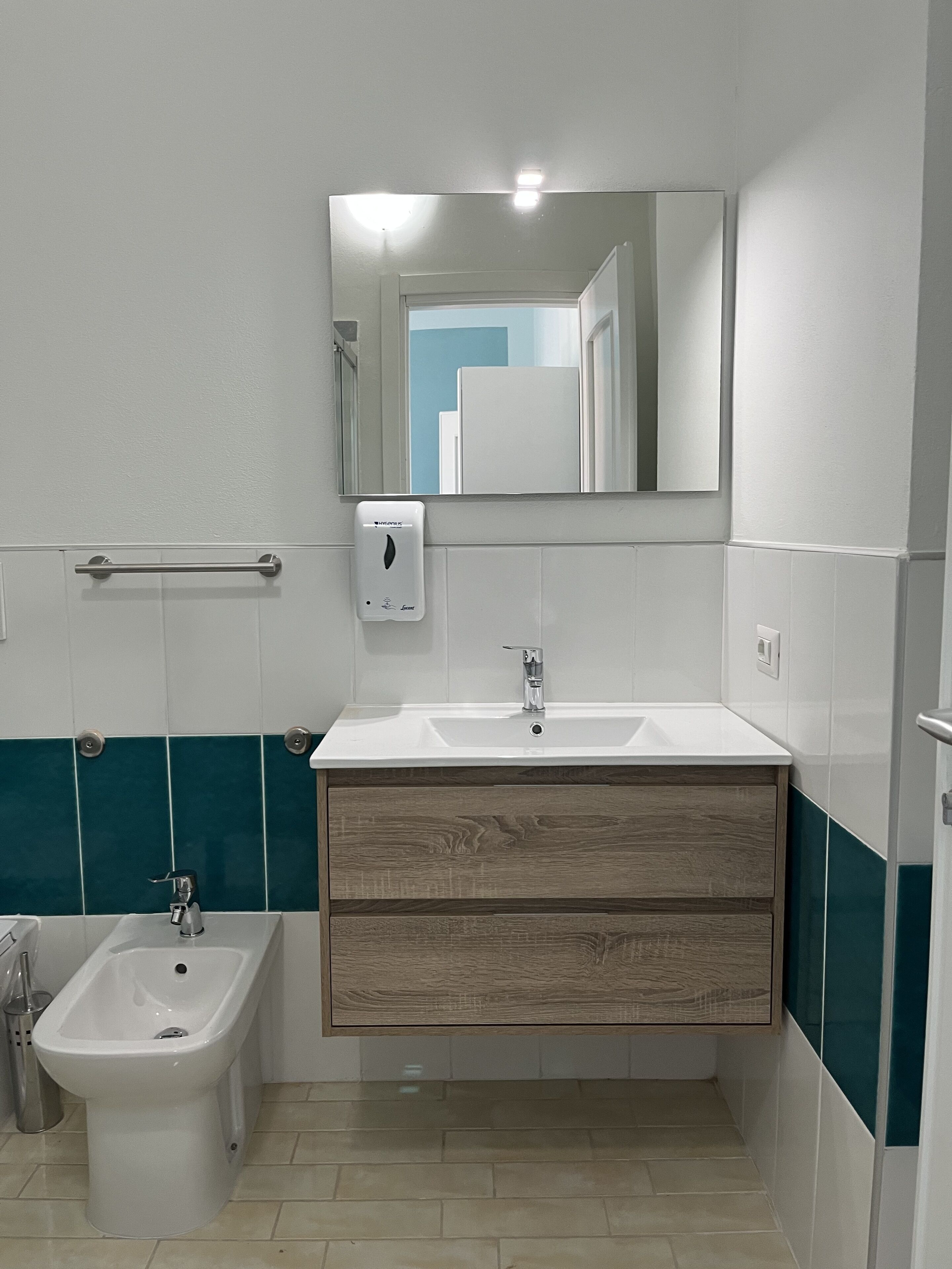 Ein Badezimmer mit Waschbecken in Weiß, einem Spiegel, einem Bidet und Fliesen in Türkis und Weiß.