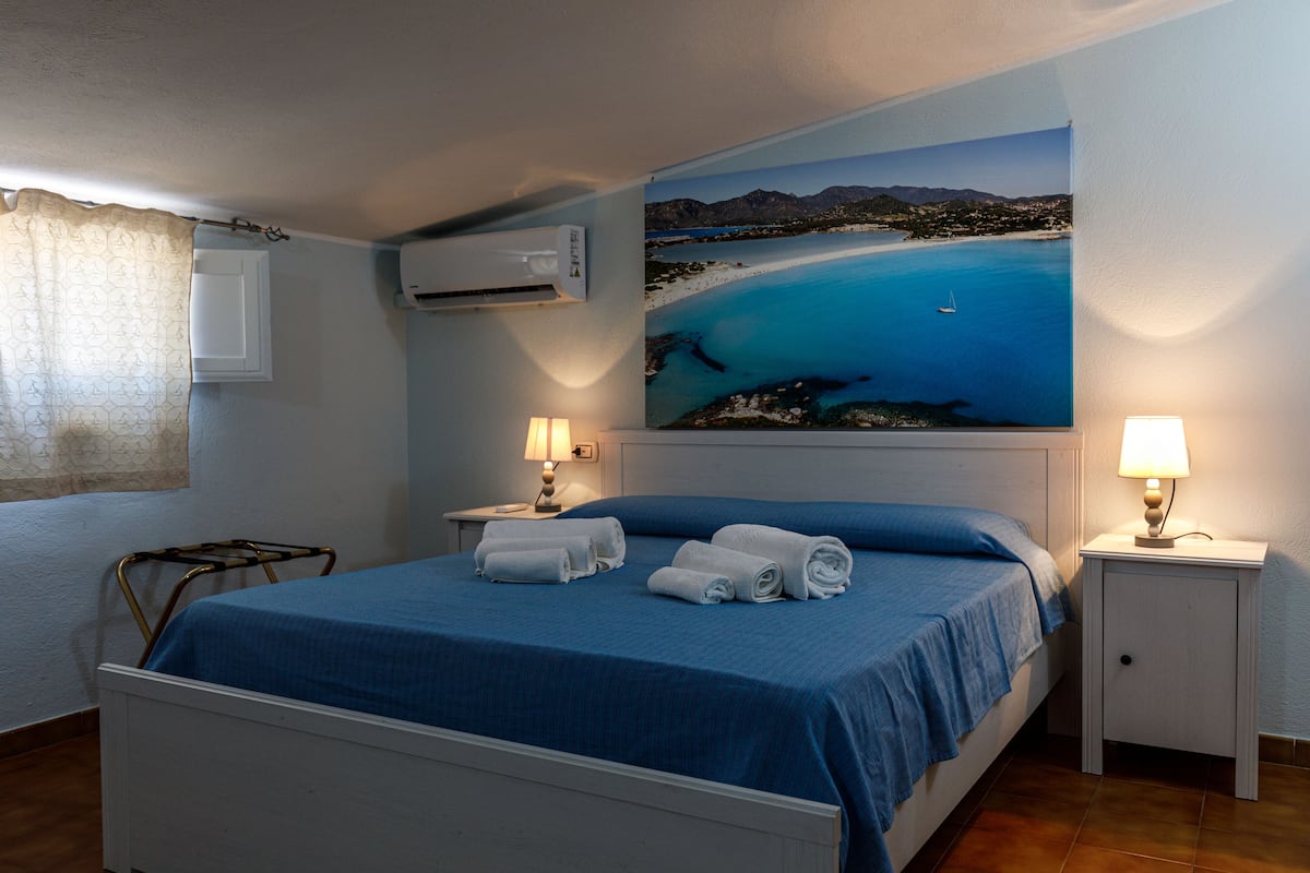 A bedroom with a bed, two bedside tables, a wall-mounted air conditioner, and a large painting of a coastal scene.