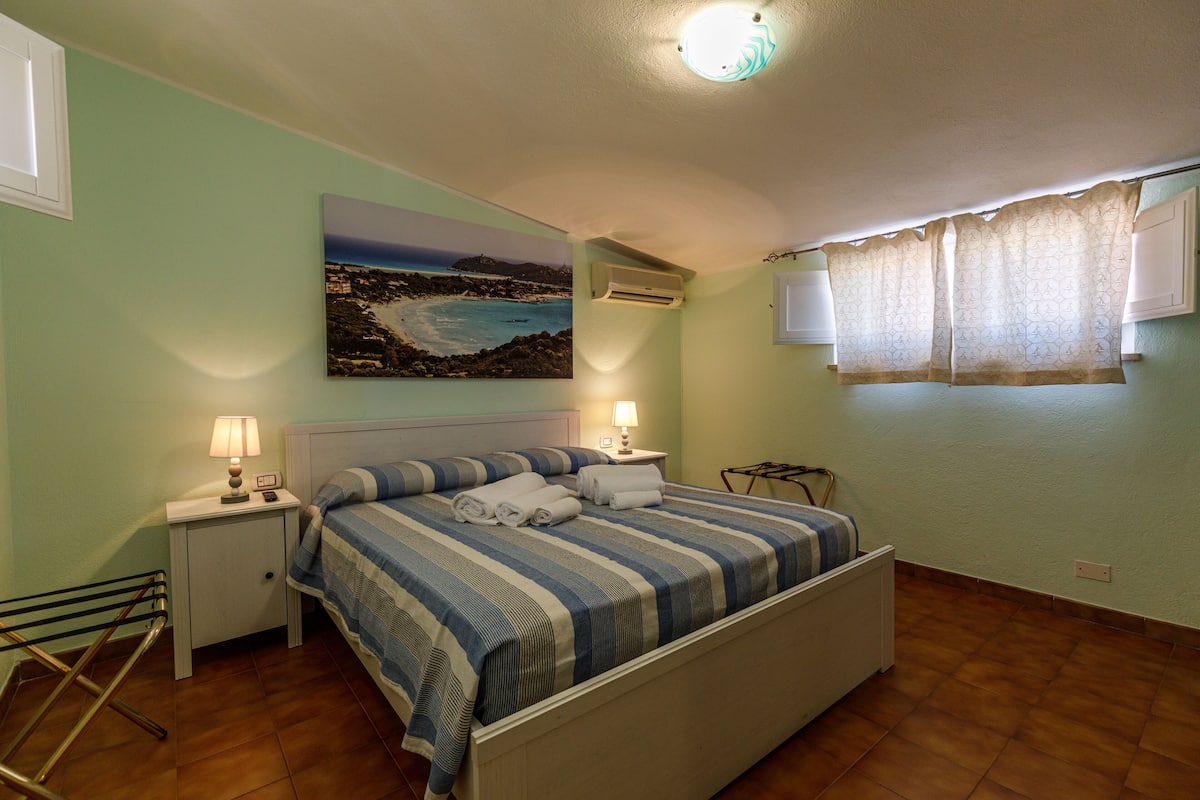 A bedroom with a bed, a nightstand with a lamp, a painting of a coastal scene, and a window with curtains.
