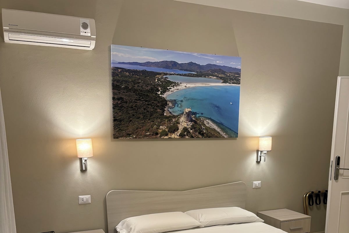 A hotel room with a bed, a nightstand, a lamp, an air conditioning unit, and a painting of a coastal landscape.