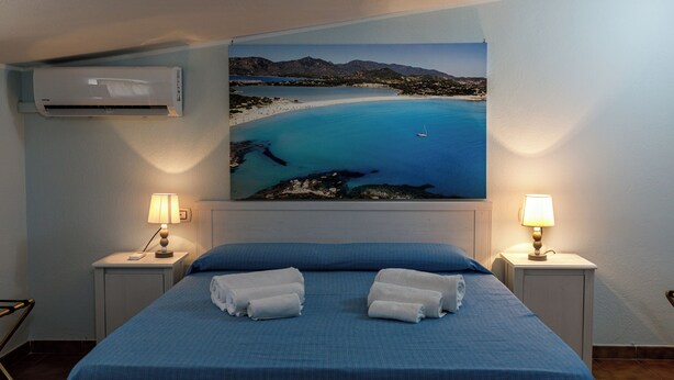 A hotel room with a bed, two bedside lamps, a painting of a coastal scene, and an air conditioning unit. - Casa Tacconi (Villasimius)