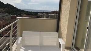 A white wicker sofa and matching ottoman on a balcony with a view of the sea and buildings. - Casa Tacconi (Villasimius)