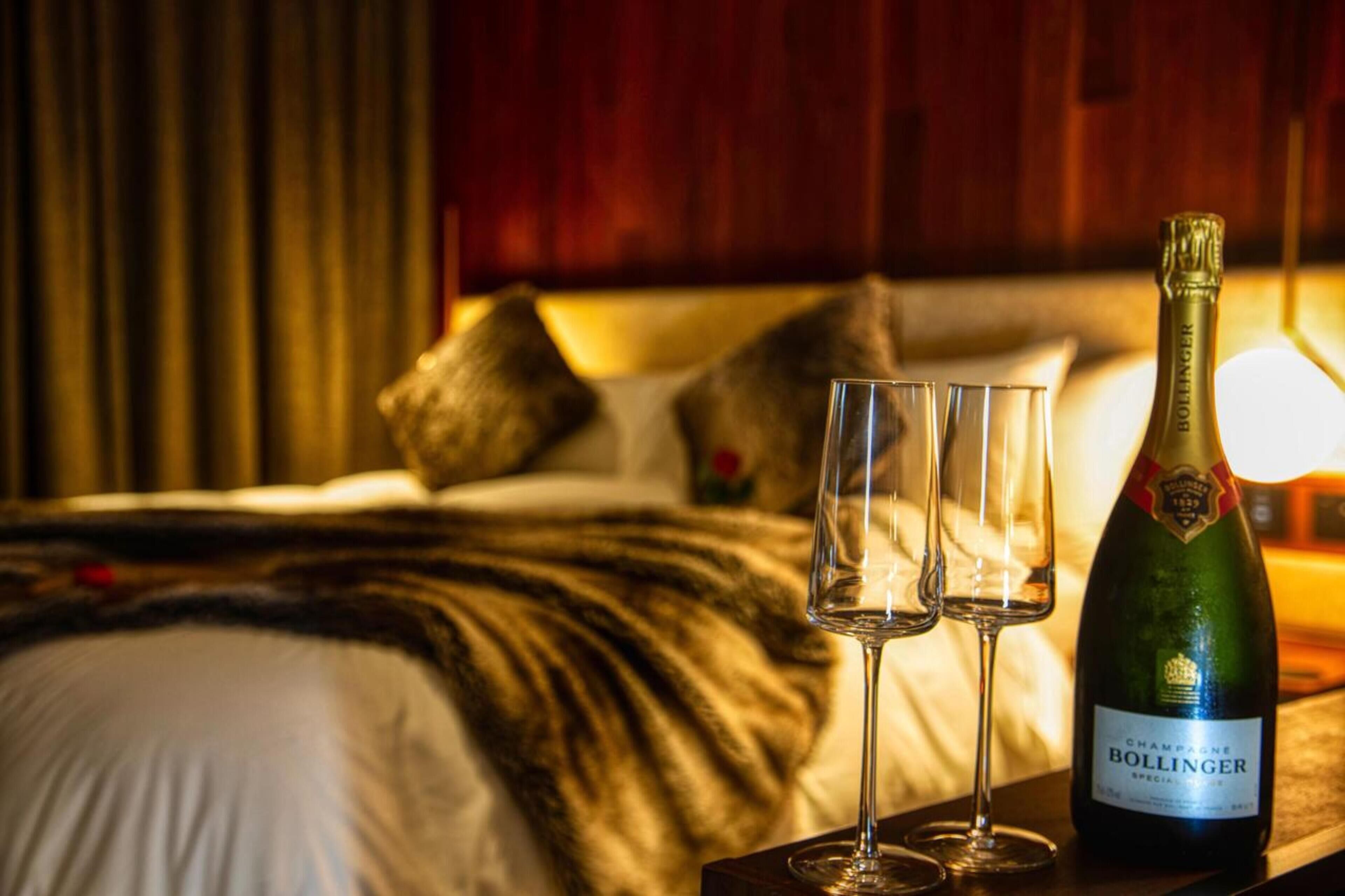 Una cama con una manta, almohadas y dos vasos vacíos sobre una mesita de noche con una botella de champán.