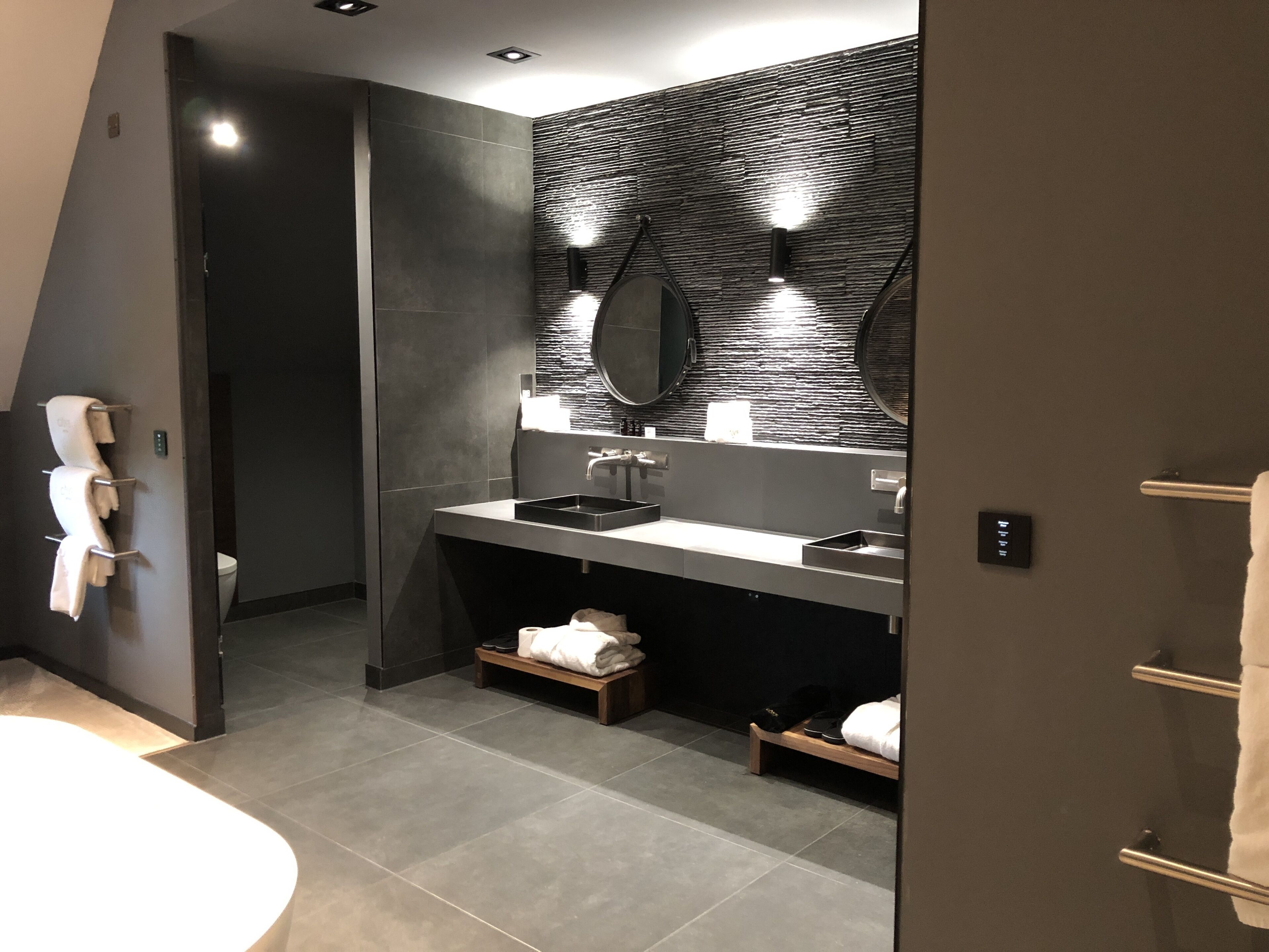 A modern bathroom with a dark tiled wall, a large mirror, and a sink with a black basin. There is a towel rack with towels and a small wooden shelf with a towel and a pair of shoes.